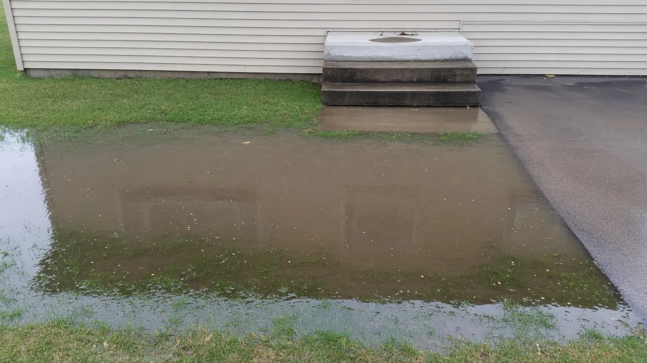 Large puddle of brown water on a green lawn beside concrete steps and a dark driveway at the side of a house.