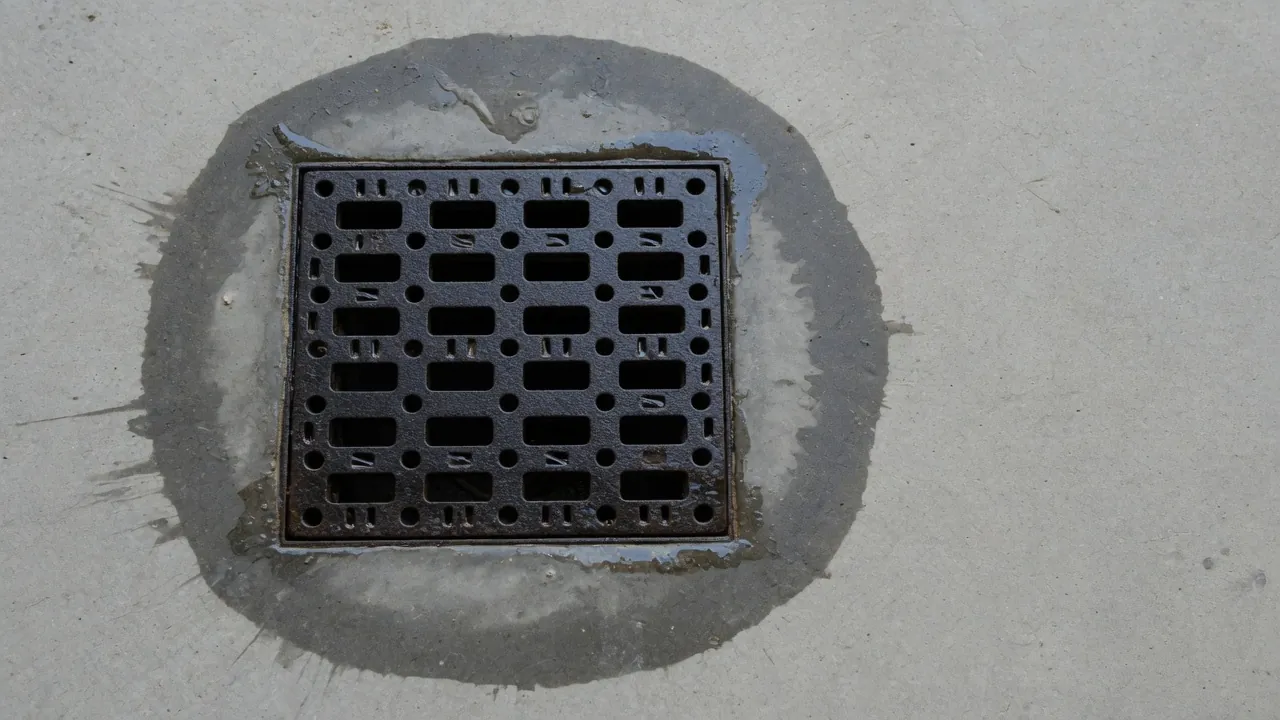 Square dark iron drain grate in a light gray concrete floor with a wide dark damp ring around it.