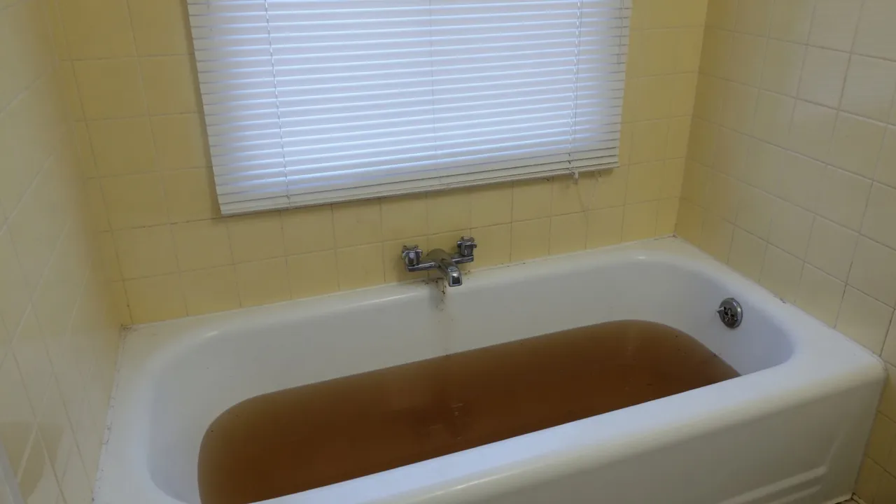 White bathtub with murky brown water, a chrome faucet and pale yellow wall tiles.