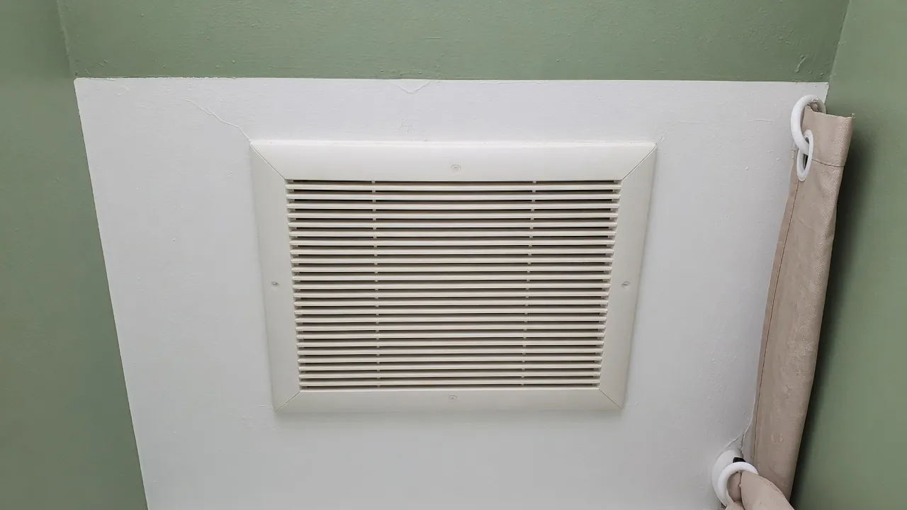 White louvered vent cover on a sage green wall beside a tan curtain.