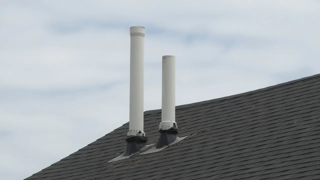 Two white plastic pipes rising through dark gray roof shingles under an overcast sky.
